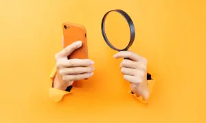 Ręce trzymające pomarańczowy iPhone i lupę, sugerujące, jak namierzyć iPhone po IMEI.