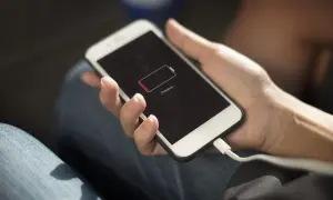 Dłoń trzyma iPhone'a z ikoną niskiego poziomu baterii. Podłączony kabel ładowania sugeruje, że szukasz sposobów, jak oszczędzić baterię w iPhone.
