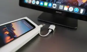 iPhone podłączony do komputera kablem, gotowy do zgrania zdjęć. Na ekranie komputera widać ikonki aplikacji.
