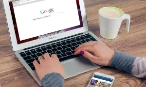 Kobieta pisze na laptopie, szukając w Google informacji, jak zmienić przeglądarkę. Obok stoi kubek kawy i telefon.