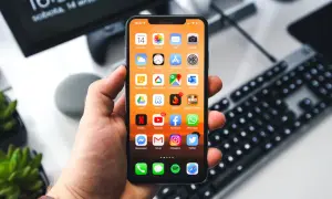 Dłoń trzyma iPhone'a z widocznymi ikonami aplikacji. Na ekranie komputera w tle widnieje godzina 16:37. Zastanawiasz się, jak wyłączyć iPhone'a?