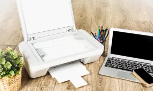 Jak podłączyć drukarkę Brother do laptopa? Na biurku drukarka, laptop, telefon i koszyk z przyborami.
