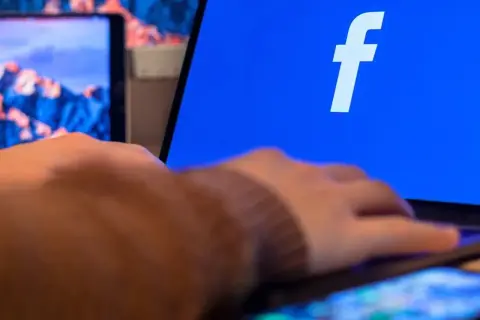 Dłoń na telefonie, obok ekranu laptopa z logo Facebooka. Dowiedz się, jak zaktualizować Facebooka na telefonie, by cieszyć się nowymi funkcjami.