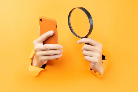 Ręce trzymające pomarańczowy iPhone i lupę, sugerujące, jak namierzyć iPhone po IMEI.