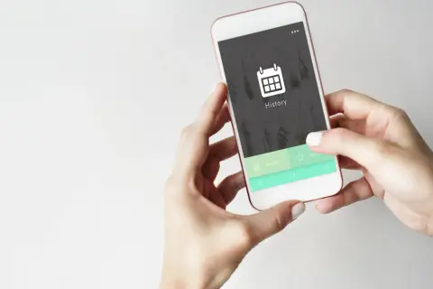 Dłonie trzymające smartfon z ikoną kalendarza i napisem "History" na ekranie.