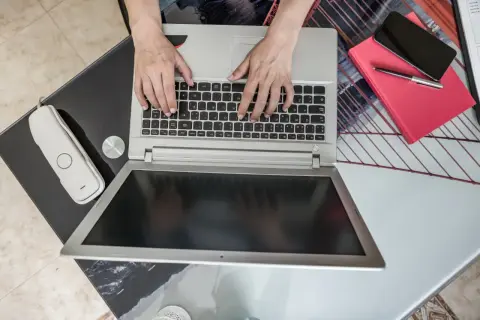 Ręce piszą na laptopie, obok telefon i czerwona książka. Zastanawiasz się, jaki laptop kupić do domu?