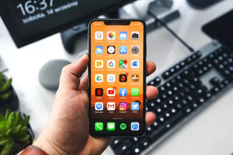 Dłoń trzyma iPhone'a z widocznymi ikonami aplikacji. Na ekranie komputera w tle widnieje godzina 16:37. Zastanawiasz się, jak wyłączyć iPhone'a?