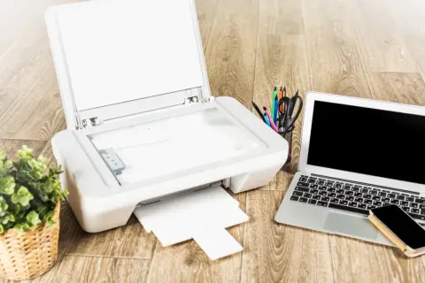 Jak podłączyć drukarkę Brother do laptopa? Na biurku drukarka, laptop, telefon i koszyk z przyborami.