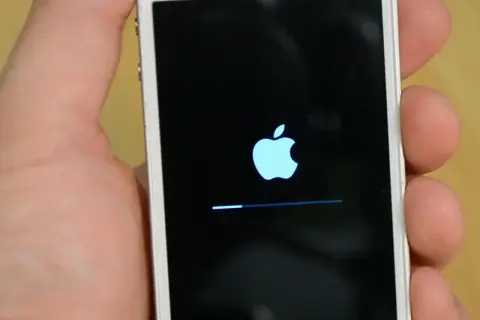 Dłoń trzyma biały iPhone z logo Apple i paskiem postępu. To ekran, który pojawia się, gdy próbujesz jak zresetować iPhone.