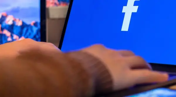 Dłoń na telefonie, obok ekranu laptopa z logo Facebooka. Dowiedz się, jak zaktualizować Facebooka na telefonie, by cieszyć się nowymi funkcjami.