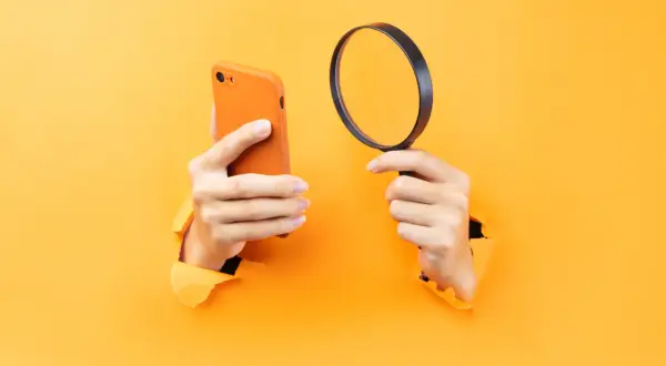 Ręce trzymające pomarańczowy iPhone i lupę, sugerujące, jak namierzyć iPhone po IMEI.