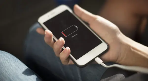 Dłoń trzyma iPhone'a z ikoną niskiego poziomu baterii. Podłączony kabel ładowania sugeruje, że szukasz sposobów, jak oszczędzić baterię w iPhone.