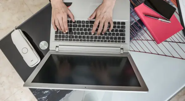 Ręce piszą na laptopie, obok telefon i czerwona książka. Zastanawiasz się, jaki laptop kupić do domu?