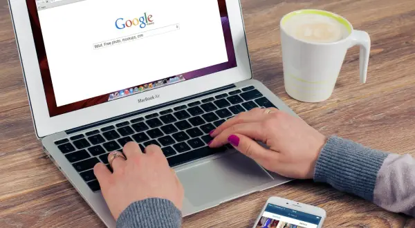 Kobieta pisze na laptopie, szukając w Google informacji, jak zmienić przeglądarkę. Obok stoi kubek kawy i telefon.