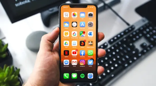 Dłoń trzyma iPhone'a z widocznymi ikonami aplikacji. Na ekranie komputera w tle widnieje godzina 16:37. Zastanawiasz się, jak wyłączyć iPhone'a?
