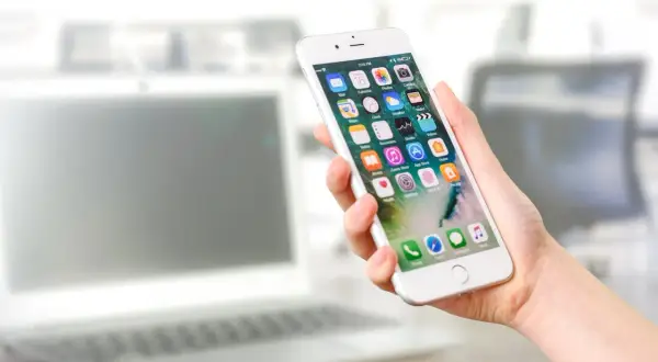 Dłoń trzyma iPhone'a z widocznymi ikonami aplikacji. Dowiedz się, jak odinstalować aplikacje, aby zwolnić miejsce.