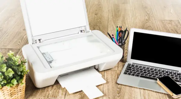Jak podłączyć drukarkę Brother do laptopa? Na biurku drukarka, laptop, telefon i koszyk z przyborami.