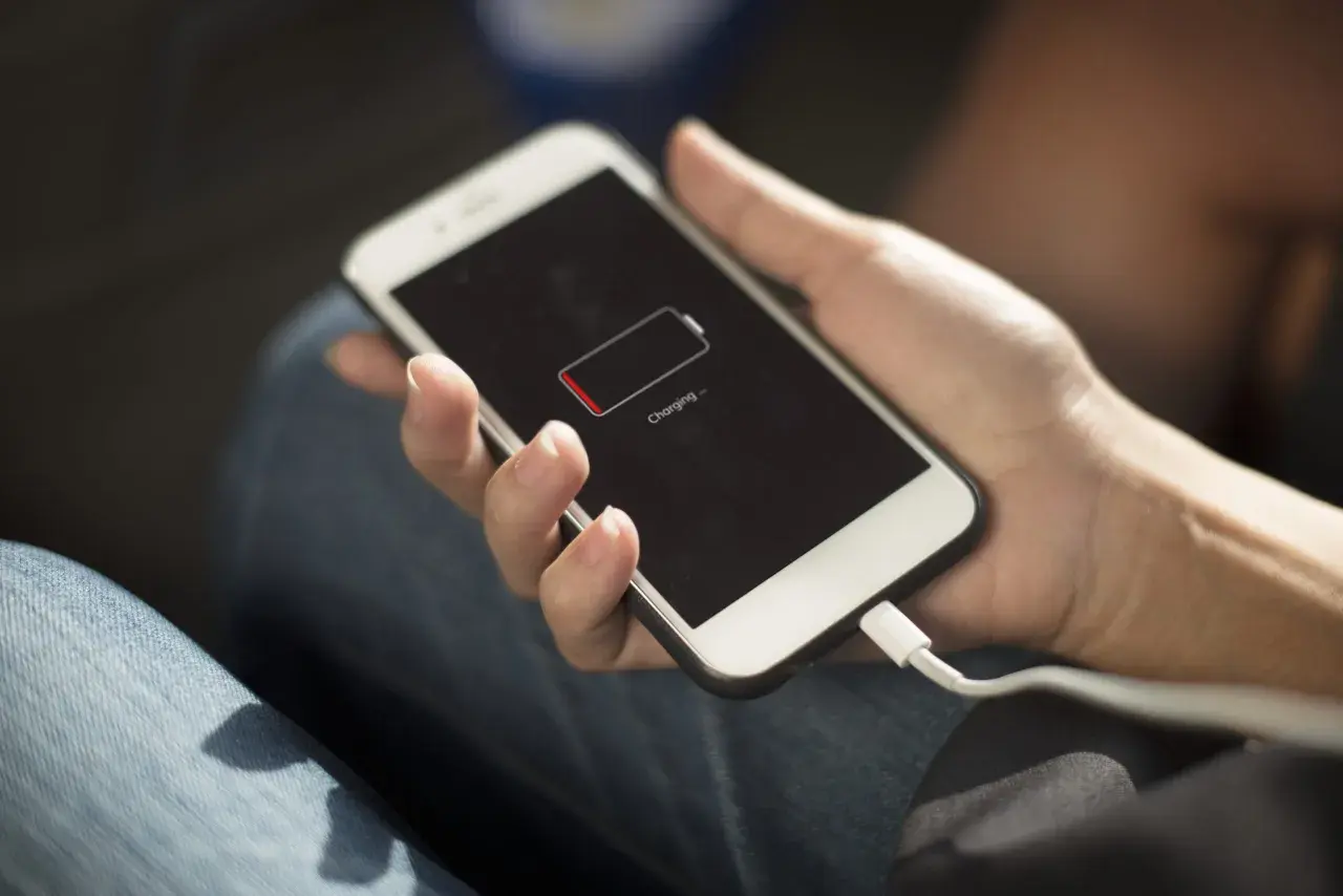 Dłoń trzyma iPhone'a z ikoną niskiego poziomu baterii. Podłączony kabel ładowania sugeruje, że szukasz sposobów, jak oszczędzić baterię w iPhone.