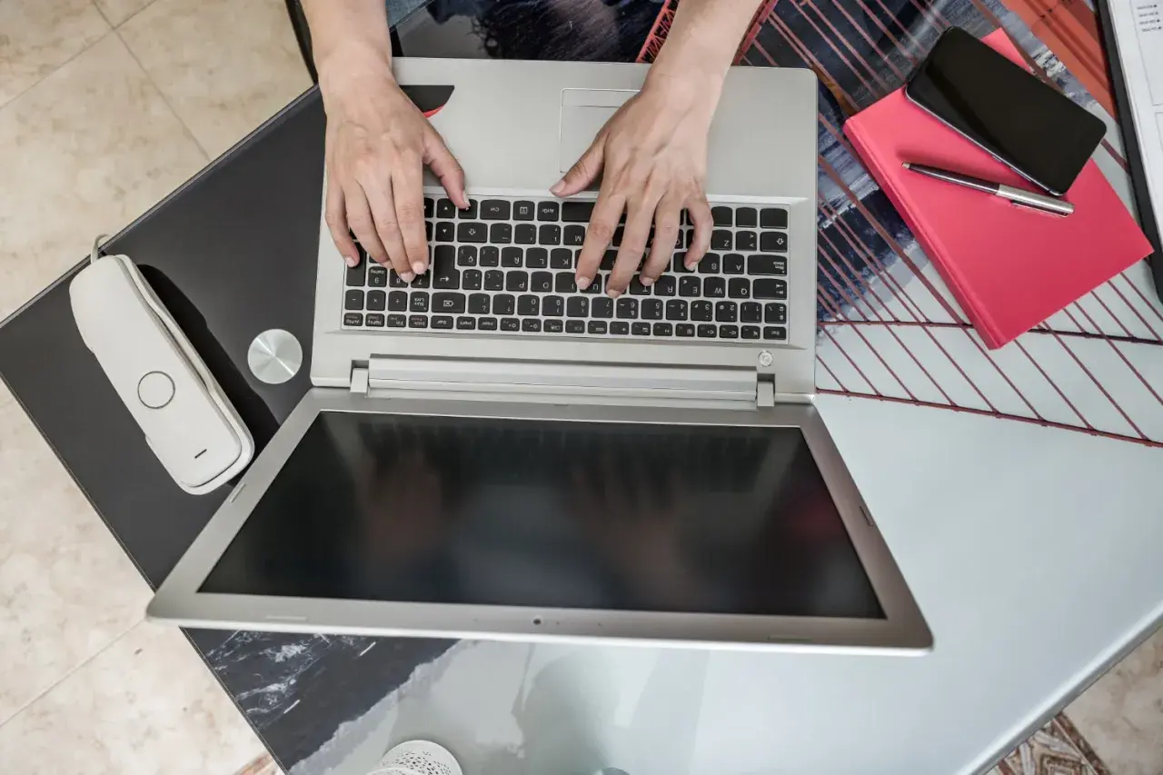 Ręce piszą na laptopie, obok telefon i czerwona książka. Zastanawiasz się, jaki laptop kupić do domu?