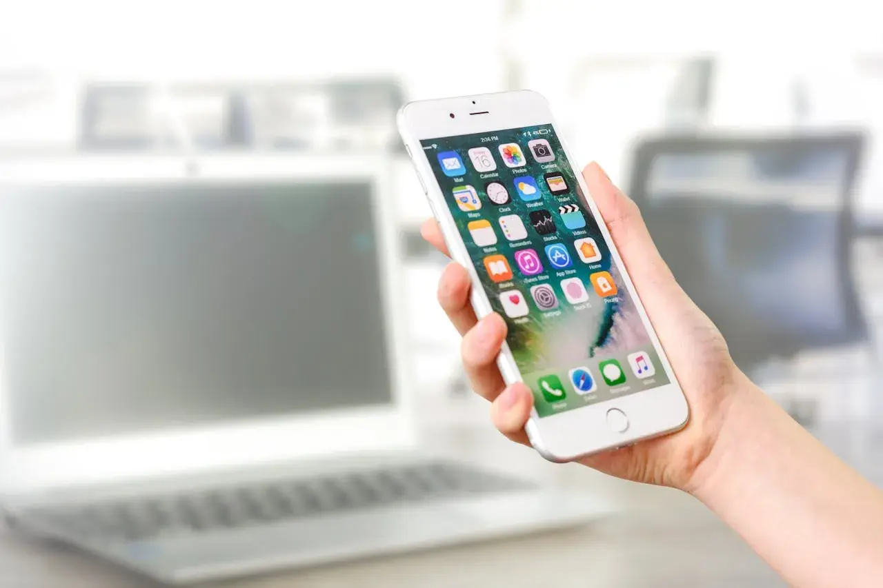 Dłoń trzyma iPhone'a z widocznymi ikonami aplikacji. Dowiedz się, jak odinstalować aplikacje, aby zwolnić miejsce.