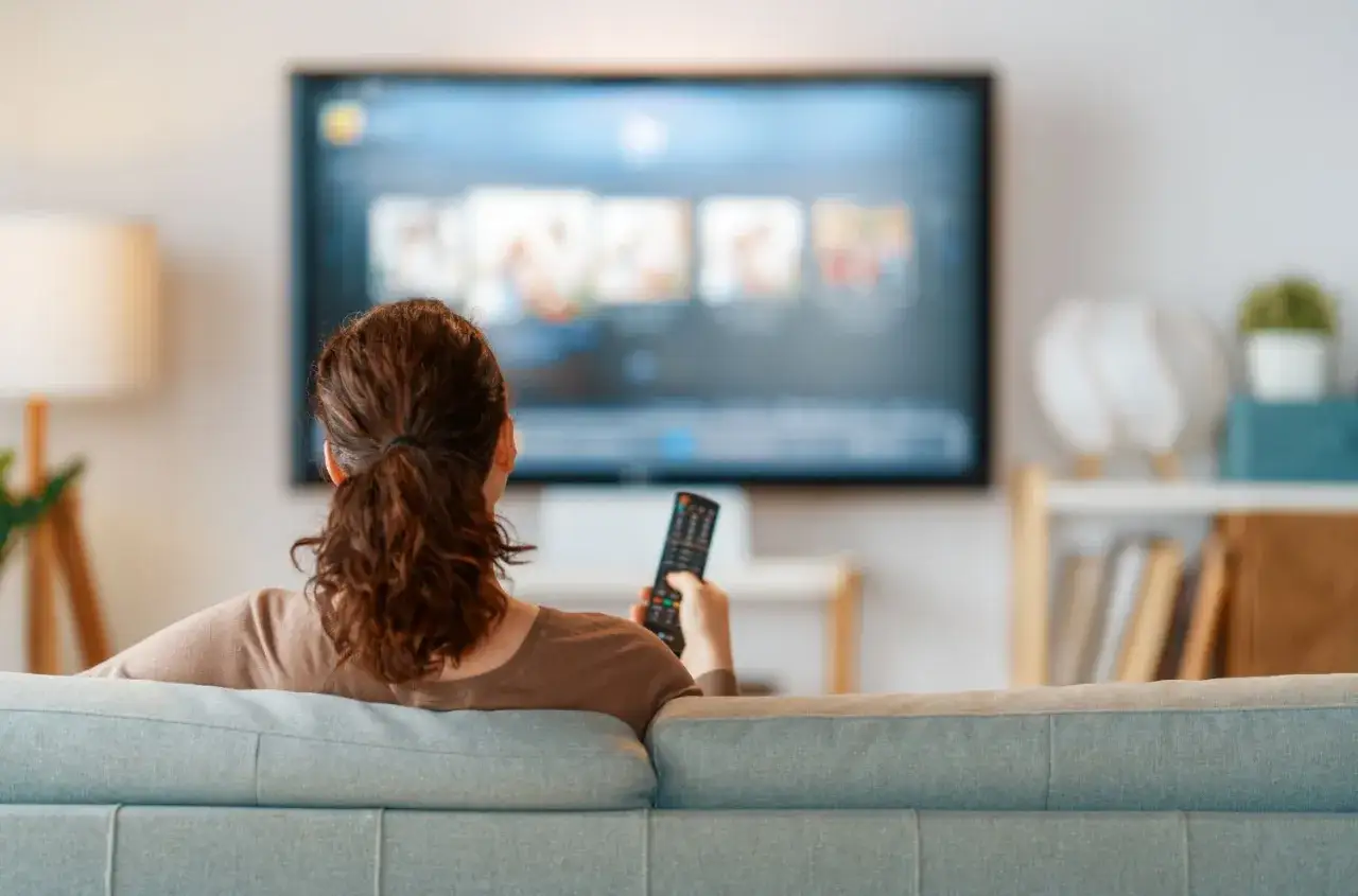 Kobieta z pilotem w ręku ogląda telewizor, zastanawiając się, jak przenieść obraz z laptopa na telewizor bezprzewodowo.